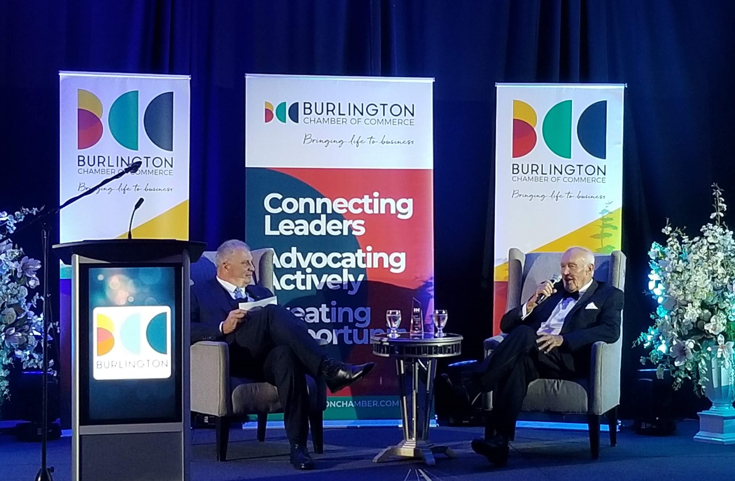 Two men sit in arm chairs in front of three standing signs. The man on the right is speaking into a microphone. The signs are printed with the Burlington Chamber of Commerce name and logo.