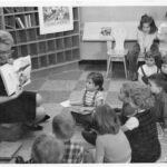 A black and white image of a woman wearing glasses sitting on a low block stool, and reading to a group of rapt children at Burlington Public Library in 1963.