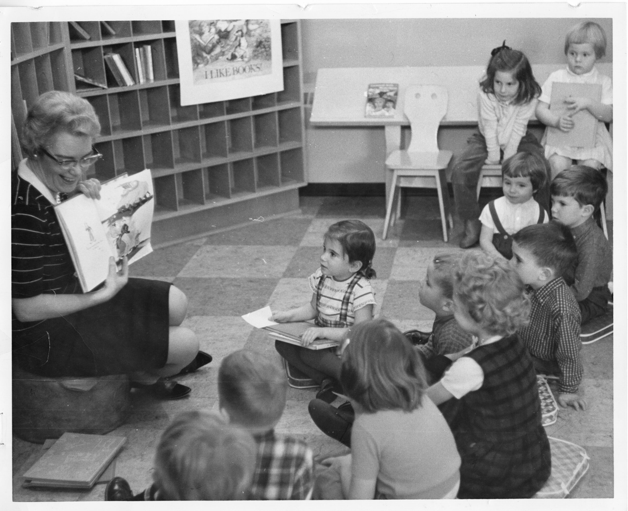 A black and white image of a woman wearing glasses sitting on a low block stool, and reading to a group of rapt children at Burlington Public Library in 1963.