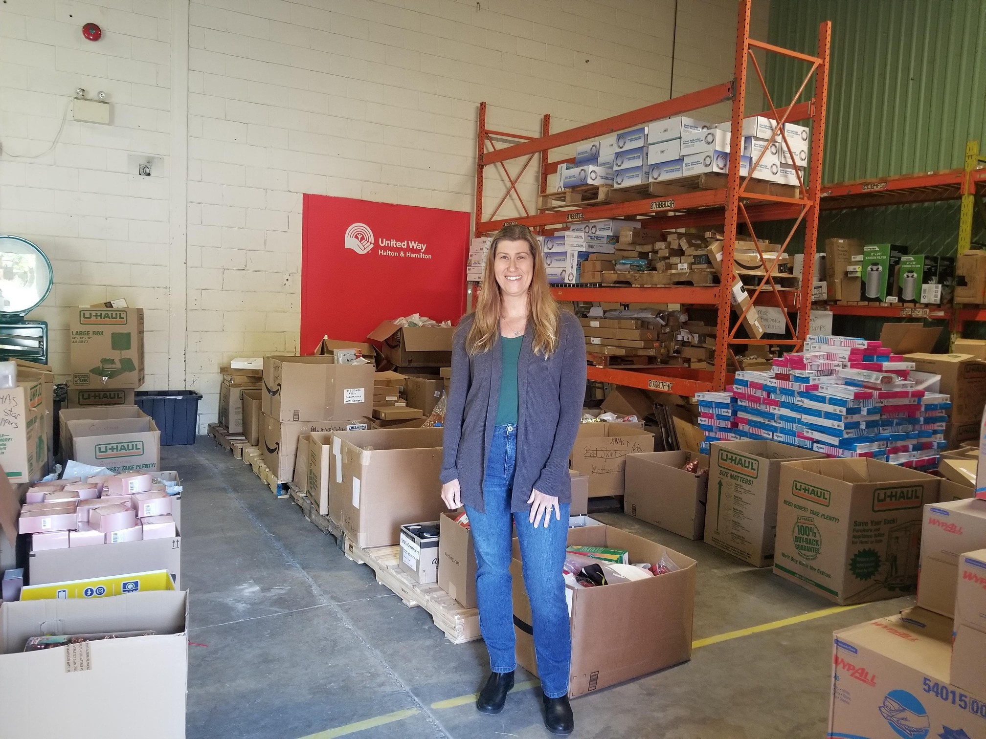 A woman stands in front of boxes and a tall shelf filled with boxes in a warehouse.