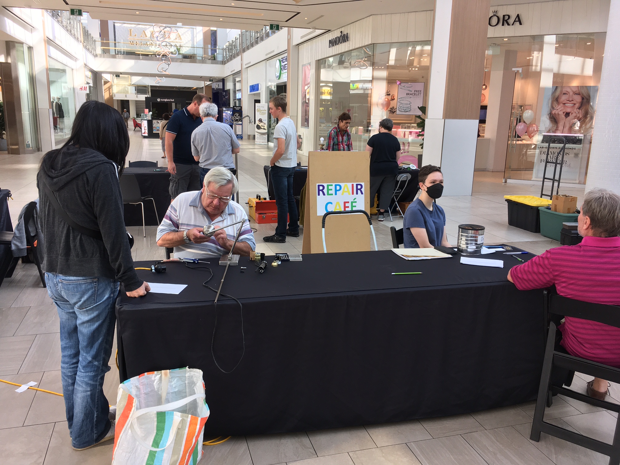 Two people at a table where to men sit, working on small appliances to fix them, at a repair cafe event in Burlington Centre mall.