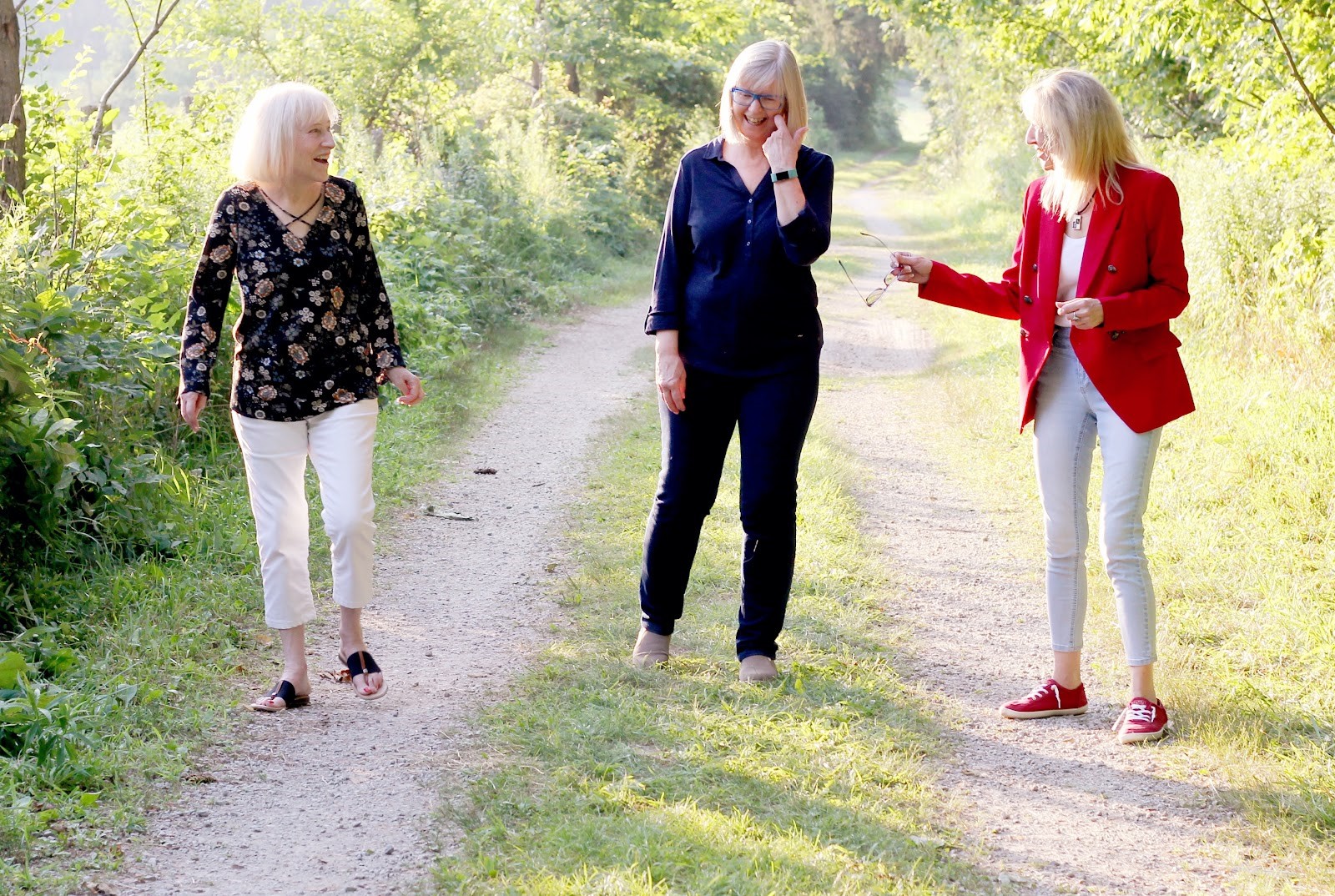Three women with white hair walk together, laughing and smiling, on a path amongst trees.