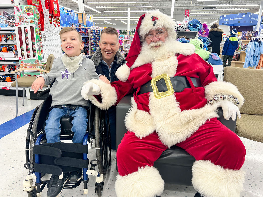 A boy in a wheelchair, posing with his dad and Santa Claus at Toys R Us Burlington.